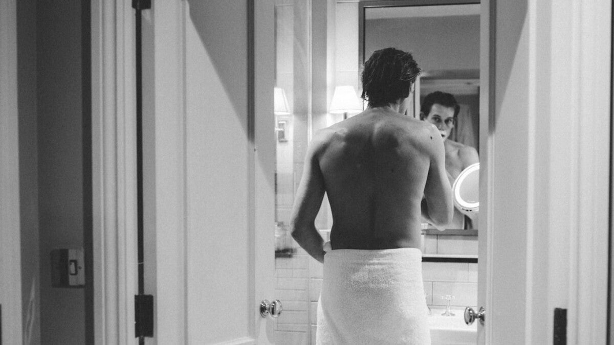 Homme séduisant appliquant un rasoir de sûreté sur son visage recouvert de mousse tout en se rasant devant un miroir de salle de bain lors d’un rasage humide traditionnel